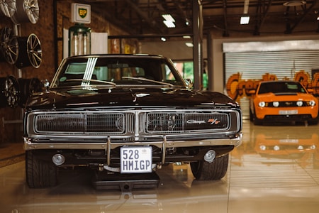A vintage black muscle car is prominently displayed in a showroom setting. Its glossy exterior and classic grille are highlighted under indoor lighting. To the left, there are several new rims mounted on the wall. In the background stands an orange modern sports car, contrasting with the classic design of the black car.