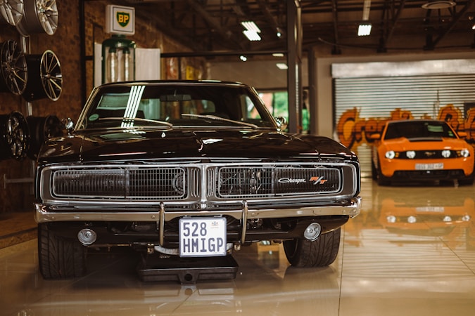 A vintage black muscle car is prominently displayed in a showroom setting. Its glossy exterior and classic grille are highlighted under indoor lighting. To the left, there are several new rims mounted on the wall. In the background stands an orange modern sports car, contrasting with the classic design of the black car.