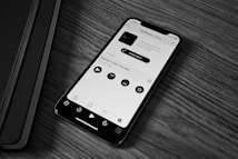 A smartphone displaying a podcast app screen is placed on a wooden surface next to a closed black notebook. The phone screen shows a podcast titled 'The Realmac Show' with controls for play, pause, and other options.