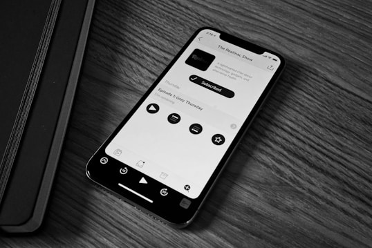 A smartphone displaying a podcast app screen is placed on a wooden surface next to a closed black notebook. The phone screen shows a podcast titled 'The Realmac Show' with controls for play, pause, and other options.