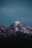 A snow-capped mountain peak set against a dark blue sky, with the soft glow of twilight accentuating the rugged textures and features of the rocky summit.