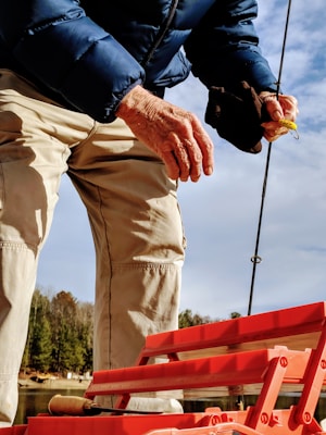 A close-up of a person preparing fishing tackle. The individual is wearing a blue puffer jacket and beige pants. The focus is on their hands, which are holding a fishing rod and a yellow lure. A red plastic tackle box is visible in the foreground. The background features a scenic view with trees and a cloudy sky.