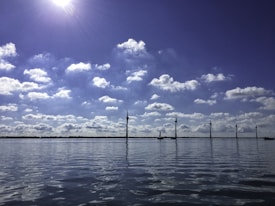 A calm body of water reflects a blue sky filled with scattered clouds. Several wind turbines stand in a line along the horizon, harnessing wind energy. A bright sun shines above, casting subtle reflections on the water's surface.