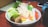 A bowl filled with a variety of fresh vegetables and tofu. There are lettuce leaves, mushrooms, enoki mushrooms, tofu cubes, and shaped vegetable slices resembling stars. The bowl sits on a wooden table with a smartphone and a teapot partially visible in the background.