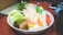 A bowl filled with a variety of fresh vegetables and tofu. There are lettuce leaves, mushrooms, enoki mushrooms, tofu cubes, and shaped vegetable slices resembling stars. The bowl sits on a wooden table with a smartphone and a teapot partially visible in the background.