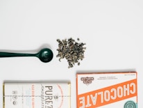 A flat lay composition featuring a green scoop, a pile of loose leaf tea or herbs, and two wrapped chocolate bars on either side. The chocolate bars have distinct vintage-style packaging with one labeled in orange and the other in white with a 'Salted Almond' descriptor.