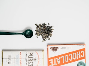 A flat lay composition featuring a green scoop, a pile of loose leaf tea or herbs, and two wrapped chocolate bars on either side. The chocolate bars have distinct vintage-style packaging with one labeled in orange and the other in white with a 'Salted Almond' descriptor.