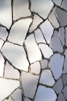A mosaic pattern composed of irregularly shaped white tiles, featuring visible grout lines in between. The tiles are arranged in a random order, creating a textured surface with various shades of white and gray under natural lighting.