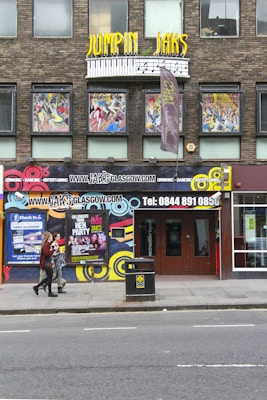 A lively street scene features the exterior of a club called Jumpin Jaks. The facade is decorated with colorful artwork and vibrant signage, including a prominent yellow sign with musical notes design. Posters advertising events and parties are displayed, and two people are walking past the entrance on the sidewalk.