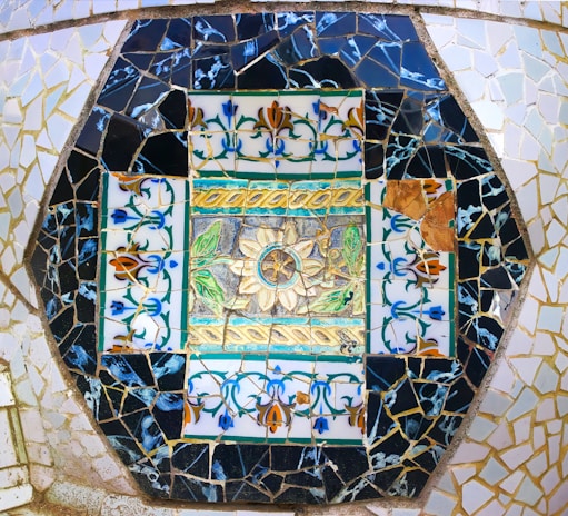 A colorful mosaic composed of intricately arranged tiles. Central to the design is a floral motif surrounded by vibrant patterns of leaves and abstract shapes. The mosaic is framed by shards of black and white tiles, creating a geometric border.