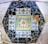 A colorful mosaic composed of intricately arranged tiles. Central to the design is a floral motif surrounded by vibrant patterns of leaves and abstract shapes. The mosaic is framed by shards of black and white tiles, creating a geometric border.