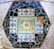 A colorful mosaic composed of intricately arranged tiles. Central to the design is a floral motif surrounded by vibrant patterns of leaves and abstract shapes. The mosaic is framed by shards of black and white tiles, creating a geometric border.