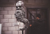 A metal sculpture featuring a skull with a long, flowing beard-like structure is prominently displayed. The sculpture has a textured, gleaming surface and stands in a room with a brick wall in the background. A person in dark clothing is seen in the background, near a door with metal accents, partially out of focus.