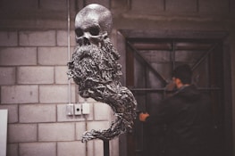 A metal sculpture featuring a skull with a long, flowing beard-like structure is prominently displayed. The sculpture has a textured, gleaming surface and stands in a room with a brick wall in the background. A person in dark clothing is seen in the background, near a door with metal accents, partially out of focus.