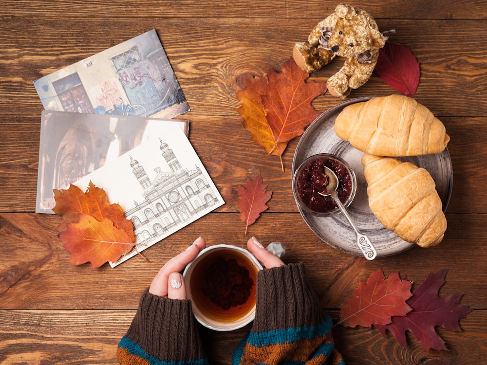 flat lay photography of person holding teacup with two croissant bread on gray tray