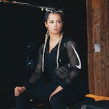 A woman with blonde braided hair is sitting on a gym bench, wearing a black zip-up hoodie with mesh sleeves and a white swoosh logo on the sleeve. She appears to be in a gym setting, indicated by the weights and gym equipment in the background. The walls are made of dark wood panels.