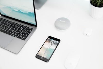 A minimalist workspace featuring a laptop with mountains as a screensaver, a smartphone displaying the time, a smart speaker, wireless earbuds, and a succulent plant. All items are neatly arranged on a white desk, creating a clean and modern aesthetic.