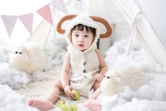 A young child is sitting on a fluffy, cloud-like surface, wearing a costume with sheep-like features, including brown horns and a woolly texture. The child appears to be in a light-colored tent setup, surrounded by plush sheep toys. The decoration includes pastel-colored flags hanging in the background.