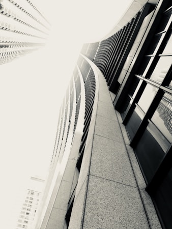 A dramatic view of tall skyscrapers emphasized by a unique angle, with the perspective drawing the eye upward along the curved lines of the building. The monochromatic color scheme adds a sense of modernity and sophistication.