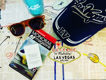 A collection of Las Vegas-themed items including a blue cap with 'Las Vegas' written on it, a pair of wooden sunglasses, a monorail magazine, and a Las Vegas souvenir cup. These items are placed on a map with iconic Las Vegas landmarks depicted on it.