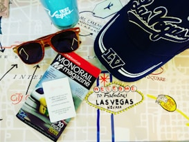 A collection of Las Vegas-themed items including a blue cap with 'Las Vegas' written on it, a pair of wooden sunglasses, a monorail magazine, and a Las Vegas souvenir cup. These items are placed on a map with iconic Las Vegas landmarks depicted on it.