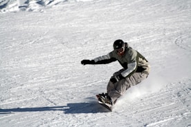 A snowboarder is skillfully maneuvering down a snow-covered slope, leaning sharply into a turn. They are dressed in a jacket and pants, wearing a helmet and goggles for safety. Snow is visibly scattering under the board, indicating speed and control.
