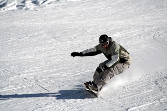A snowboarder is skillfully maneuvering down a snow-covered slope, leaning sharply into a turn. They are dressed in a jacket and pants, wearing a helmet and goggles for safety. Snow is visibly scattering under the board, indicating speed and control.