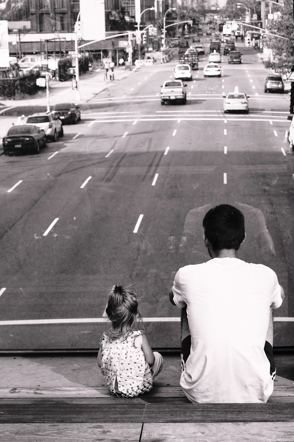 grayscale photography of man and girl sitting on staircase fronting road with passing cars