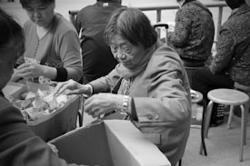 A group of people is engaged in sorting items into cardboard boxes. An elderly person wearing a jacket appears to be focused on the task, with others working collaboratively around a table filled with packages. The scene captures a moment of teamwork and concentration in an outdoor setting.