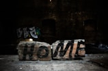 Two bricks on a surface feature the word 'HOME' spray-painted in bold black letters. The background is dark, indicating a possible abandoned building or industrial setting, with some graffiti visible on the walls.