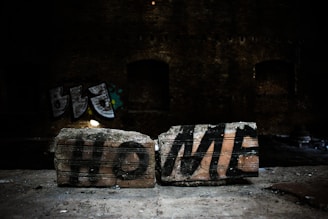 Two bricks on a surface feature the word 'HOME' spray-painted in bold black letters. The background is dark, indicating a possible abandoned building or industrial setting, with some graffiti visible on the walls.