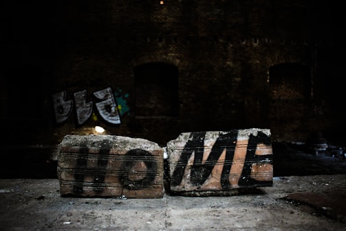 Two bricks on a surface feature the word 'HOME' spray-painted in bold black letters. The background is dark, indicating a possible abandoned building or industrial setting, with some graffiti visible on the walls.