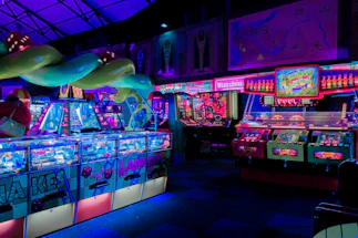 An indoor arcade filled with vibrant gaming machines lit with neon blue and pink lights. The scene includes various arcade games with flashing screens and colorful displays, and a large overhead structure resembling a green snake. The background shows illuminated signs and themed decor, contributing to an immersive entertainment atmosphere.