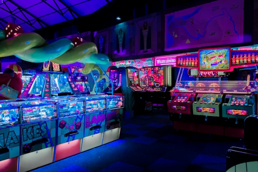 An indoor arcade filled with vibrant gaming machines lit with neon blue and pink lights. The scene includes various arcade games with flashing screens and colorful displays, and a large overhead structure resembling a green snake. The background shows illuminated signs and themed decor, contributing to an immersive entertainment atmosphere.