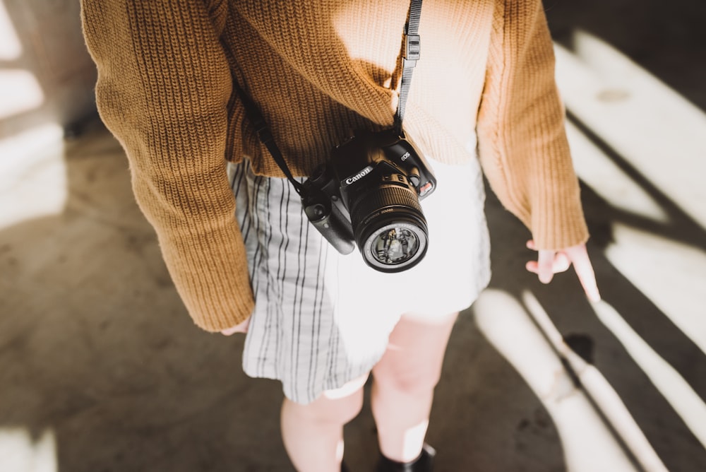 donna che indossa un maglione marrone e pantaloncini bianchi con un'imbracatura nera per fotocamera reflex digitale Canon sul suo corpo