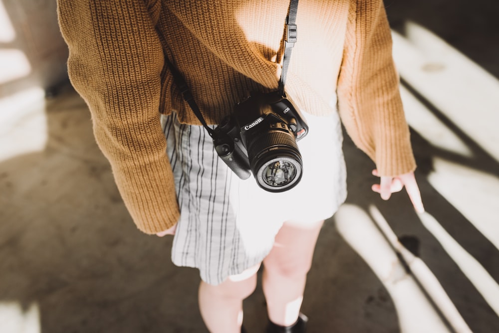 mujer con suéter marrón y pantalones cortos blancos con cabestrillo de cámara Canon DSLR negro en su cuerpo