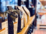 Several bags of organic wholemeal pasta are lined up on a wooden shelf in what appears to be a store. The bags display various labels with text, and the background features a blurred, softly lit setting with other shop products visible.