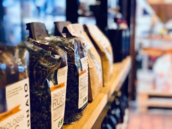 Several bags of organic wholemeal pasta are lined up on a wooden shelf in what appears to be a store. The bags display various labels with text, and the background features a blurred, softly lit setting with other shop products visible.