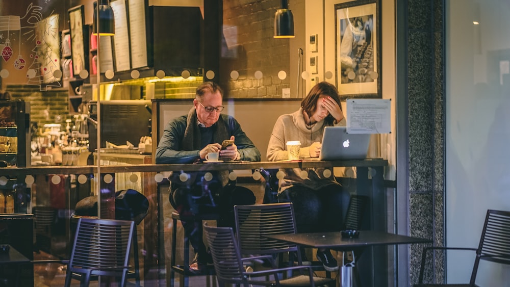 man holding smartphone sitting beside woman in front of silver MacBook in restaurant
