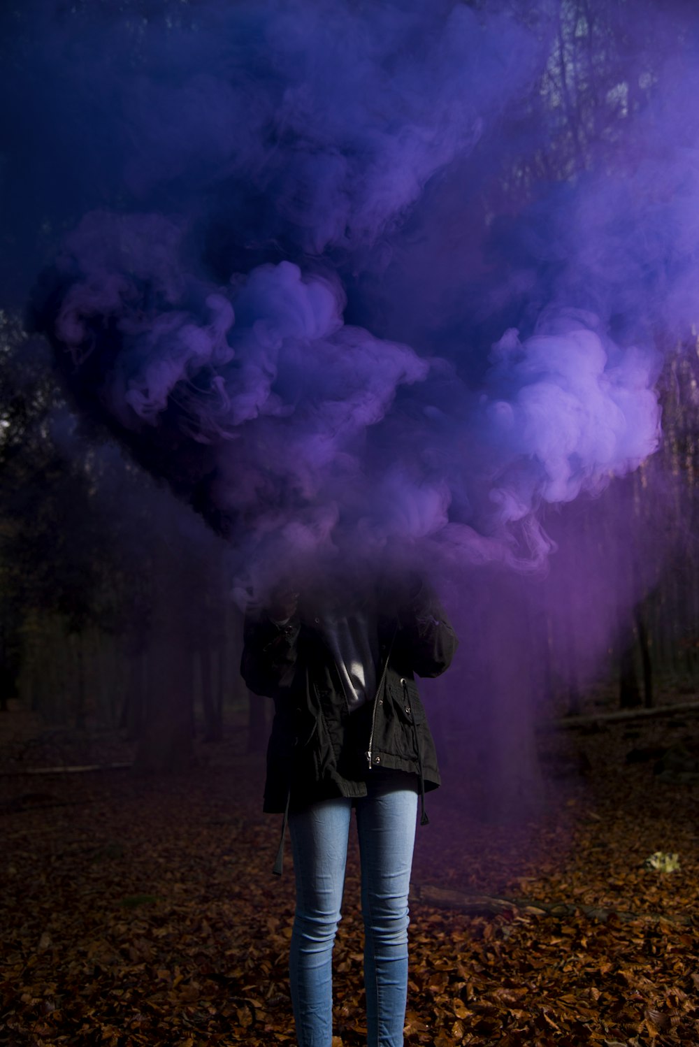 Persona con chaqueta negra y jeans azules sosteniendo una bomba de humo púrpura de pie sobre hojas secas de árboles