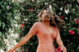 A woman with long, flowing hair is in motion, standing amidst lush green foliage with vibrant pink flowers. She is wearing a peach-colored dress, giving a sense of movement and vitality.