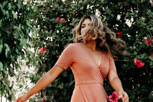 A woman with long, flowing hair is in motion, standing amidst lush green foliage with vibrant pink flowers. She is wearing a peach-colored dress, giving a sense of movement and vitality.