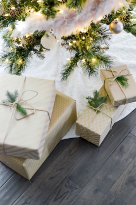 Several neatly wrapped gifts are placed under a Christmas tree, decorated with lights, pine cones, and baubles. The presents feature elegant gold and white paper with simple twine and greenery accents.