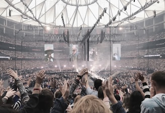 A large indoor concert venue filled with a cheering crowd. The stage is set in the center with multiple large screens displaying performers. Bright spotlights illuminate the stage area, and the audience members have their hands raised in excitement. The ceiling features an intricate pattern of beams and lights.