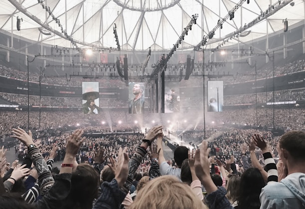 A large indoor concert venue filled with a cheering crowd. The stage is set in the center with multiple large screens displaying performers. Bright spotlights illuminate the stage area, and the audience members have their hands raised in excitement. The ceiling features an intricate pattern of beams and lights.