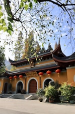 A traditional Chinese building with ornate curved roofs and red lanterns hanging across the entrance. The structure is surrounded by greenery, including branches with leaves in the foreground and trees in the background. The sunlight casts warm tones on the yellow and red colors of the building.