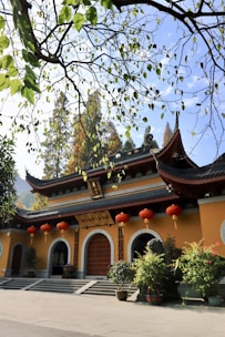 A traditional Chinese building with ornate curved roofs and red lanterns hanging across the entrance. The structure is surrounded by greenery, including branches with leaves in the foreground and trees in the background. The sunlight casts warm tones on the yellow and red colors of the building.