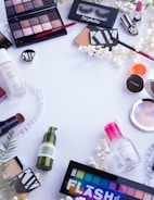 A collection of various makeup and skincare products arranged on a white surface. The items include eyeshadow palettes, false eyelashes, brushes, lipsticks, foundation, eyelash curler, and other cosmetics. There are also decorative white flowers scattered around the products.