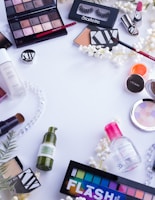 A collection of various makeup and skincare products arranged on a white surface. The items include eyeshadow palettes, false eyelashes, brushes, lipsticks, foundation, eyelash curler, and other cosmetics. There are also decorative white flowers scattered around the products.
