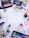 A collection of various makeup and skincare products arranged on a white surface. The items include eyeshadow palettes, false eyelashes, brushes, lipsticks, foundation, eyelash curler, and other cosmetics. There are also decorative white flowers scattered around the products.